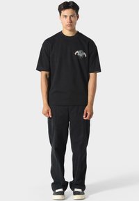 Sort bomulds t-shirt med grafisk design på brystet, parret med løstsiddende sorte bukser og mørke sneakers med hvide detaljer.