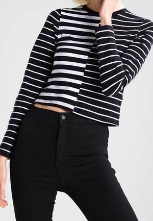 Femme portant un top court rayé noir et blanc à manches longues associé à un pantalon noir taille haute sur un fond uni.