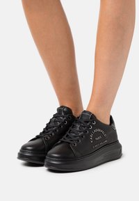 Black leather sneakers with round toe, lace-up design, padded collar, and embossed logo detailing on the side. Flat rubber sole.