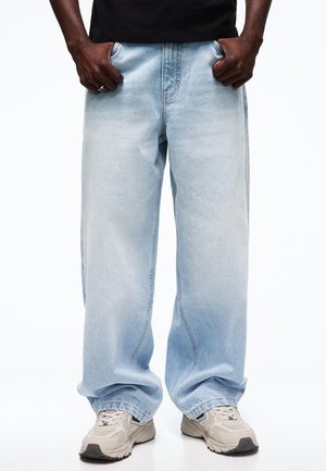 Person iført lyseblå vidde bukser, beige og hvide sneakers og en sort skjorte med hænderne i lommerne.