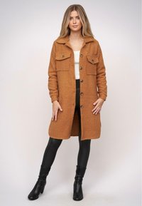 Barna, textúrált, hosszú kabát, gallérral, gombos záródással és két elülső zsebbel, fekete leggings-szel és térd középig érő fekete csizmával párosítva.