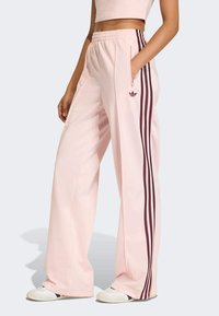 Jasnoróżowe spodnie dresowe z bocznymi kieszeniami, elastycznym pasem i trzema bordowymi paskami na każdej nogawce. Dyskretne logo na lewej stronie biodra.