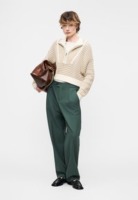 Άτομο που φοράει κρεμ πουλόβερ πλέξης με φερμουάρ στο μισό, σκούρο πράσινο παντελόνι με φαρδιά πόδια, μαύρα μοκασίνια και κρατάει καφέ δερμάτινη τσάντα με χρυσή αλυσίδα.