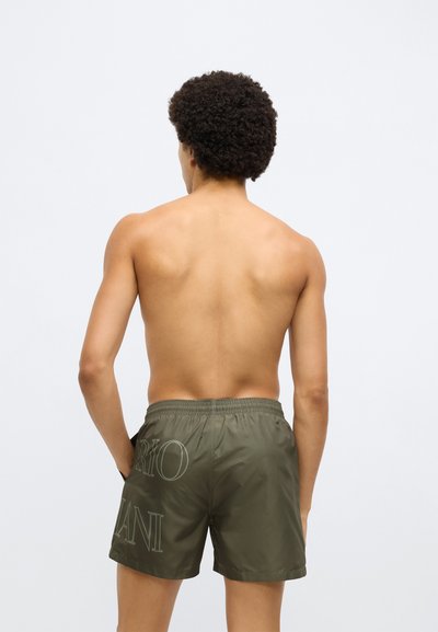 Olívazöld fürdőnadrág elasztikus derékrésszel, világos színű felirattal a hátsó részen, sima, szintetikus anyagból.