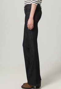 Ciemnoniebieskie dżinsy o rozszerzanym kroju nogawek, z pośrednim stanem i subtelnymi detalami przeszycia. Noszone z brązowymi botkami na obcasie.