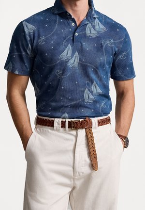 Mężczyzna w granatowej koszulce polo z nadrukiem żaglówki i kotwicy, beżowe spodnie, brązowy pleciony skórzany pasek i zegarek na nadgarstku, ręce w kieszeniach.