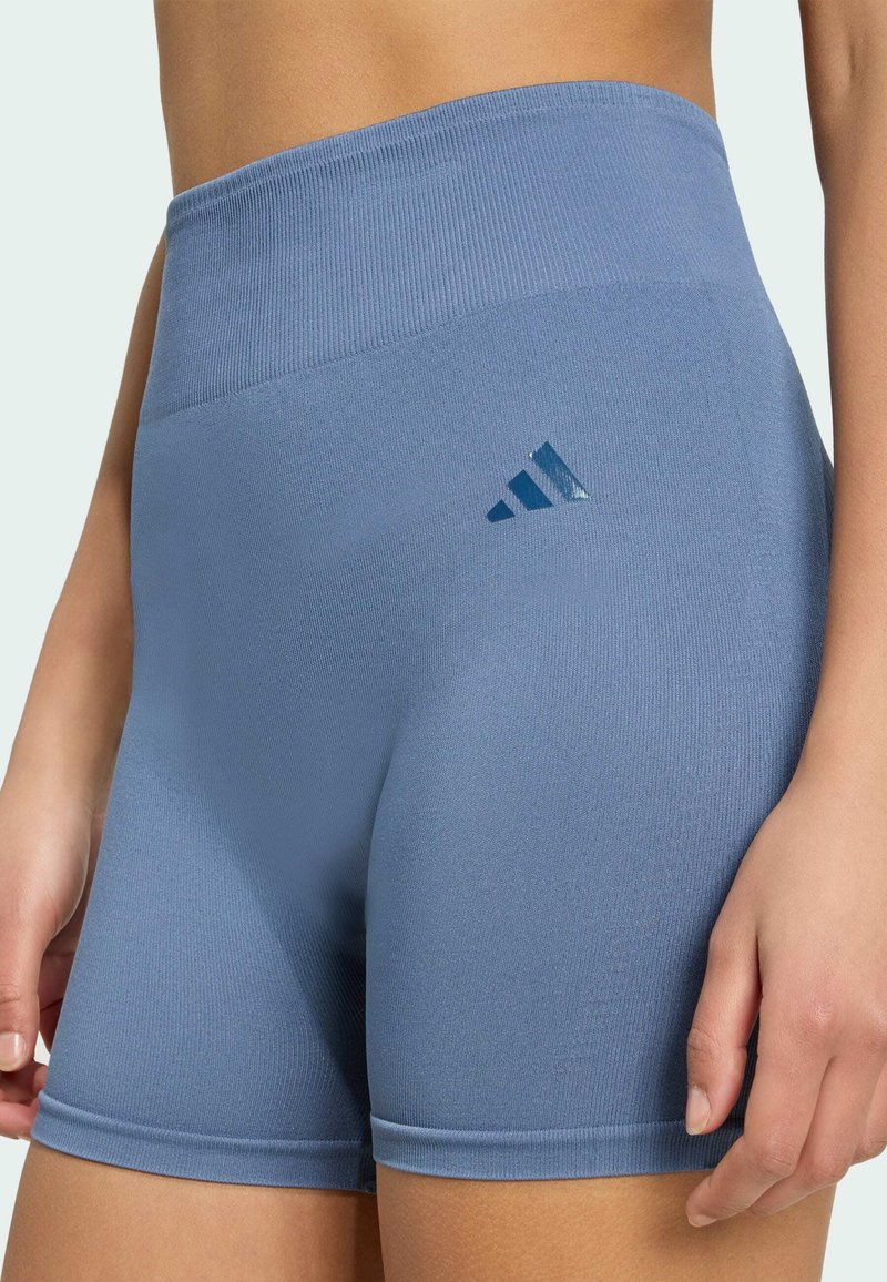 Shorts de sport bleus taille haute avec une ceinture côtelée et le logo Adidas sur la cuisse gauche, portés par une personne debout, bras détendus.