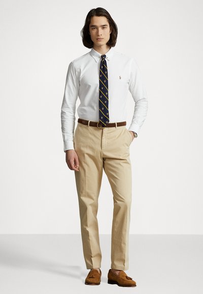 Witte overhemdd, marineblauwe stropdas met gouden accenten, beige broek, leren bruine riem en bruine suède instappers, staand tegen een neutrale achtergrond.