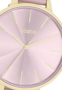 Montre ronde avec un cadran rose brossé, des index horaires en or et un boîtier doré. Elle possède une texture lisse et un design minimaliste.