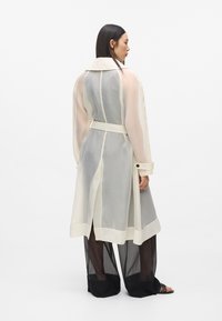 Long manteau beige transparent avec une ceinture à la taille, manches opaques contrastantes et col classique, associé à un pantalon noir large en maille.