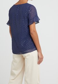 Blusa blu navy con maniche corte a balze, texture con piccoli punti dorati, dotata di scollo rotondo e materiale semitrasparente.