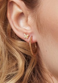 Złota kolczyk w kształcie obrączki oraz mały kolczyk w kształcie motylka z drobnymi żółtymi i przezroczystymi detalami; gładka tekstura, zbliżenie na ucho.