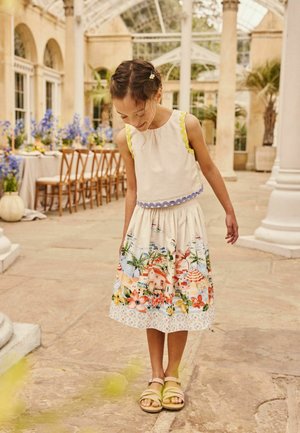 Young girl in a colorful skirt with seaside and floral prints and a sleeveless top looks down while standing in a bright indoor courtyard.