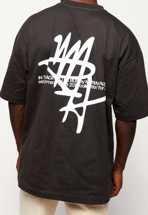 Personne portant un t-shirt noir avec un grand motif abstrait blanc et un texte blanc partiellement visible dans le dos, associé à un pantalon clair.