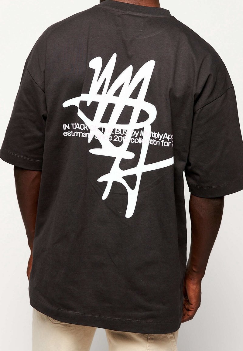 Personne portant un t-shirt noir avec un grand motif abstrait blanc et un texte blanc partiellement visible dans le dos, associé à un pantalon clair.