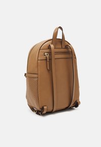 Sac à dos en cuir marron avec une fermeture éclair dorée, des poches latérales perforées, des bretelles réglables et une poignée supérieure sur fond blanc.