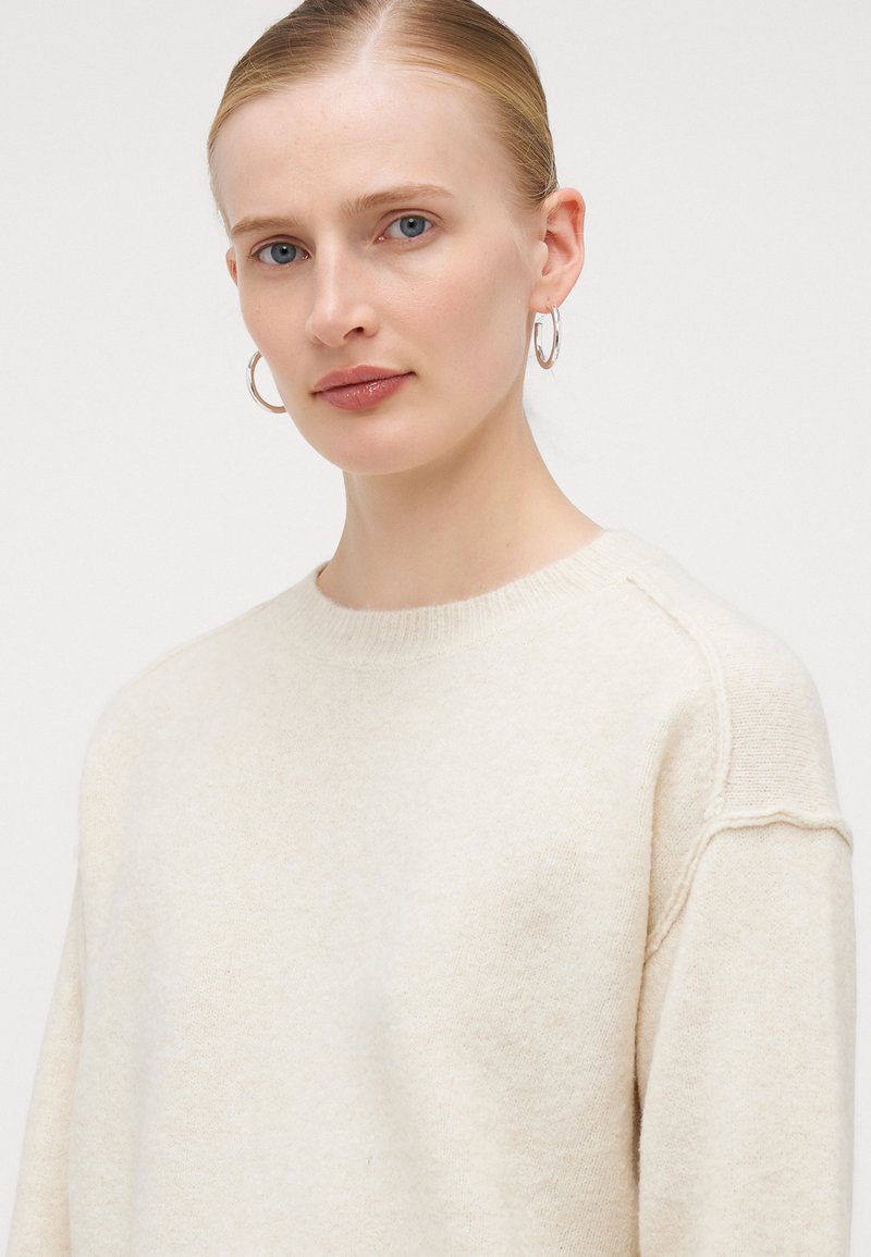 Pull en crème avec un col rond, texture douce et poignets côtelés, présentant une coupe légèrement décontractée et un minimum de détails de couture.