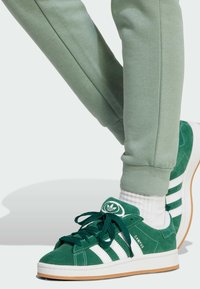 Green suede sneakers with white stripes and branding, paired with light green sweatpants and white socks, showcasing a casual look.