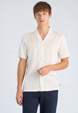 Giovane uomo con capelli castano chiaro che indossa una camicia bianca a maniche corte con bottoni e pantaloni blu navy, in piedi contro uno sfondo chiaro e uniforme.