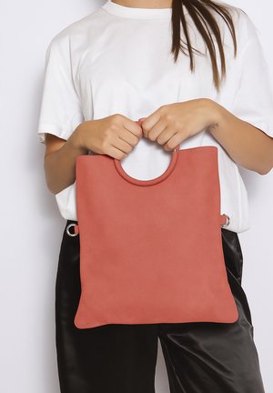 Persoon houdt een grote vierkante, roestkleurige handtas met ronde handgrepen vast, gekleed in een wit overhemd en zwarte broek.