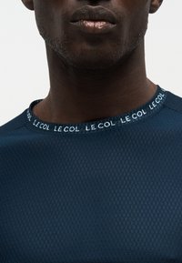 Nahaufnahme eines Mannes, der ein dunkelblaues, strukturiertes Hemd mit dem wiederholten Schriftzug „LE COL“ am Kragen trägt und dabei sein unteres Gesicht sowie seinen Hals zeigt.