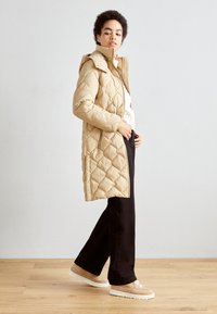 Abrigo de plumas acolchado beige con cuello alto y capucha, presenta bolsillos laterales, combinado con pantalones negros y zapatillas color beige claro.