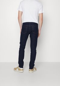 Jeans de mezclilla azul oscuro con un diseño ajustado, que cuentan con bolsillos traseros y detalles sutiles de costura; combinados con una camiseta blanca y zapatillas beige.