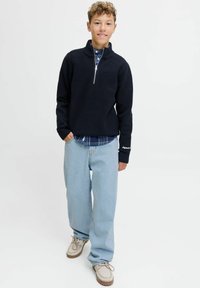 Marineblauwe wollen trui met ritssluiting, geribde manchetten, gedragen over een blauw geruite blouse. Lichtblauwe losse jeans en beige schoenen maken de outfit compleet.