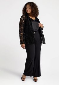 Svart spetskofta över en satin svart topp, kombinerad med högmidjade svarta byxor. Spetsen har blommönster som ger en texturerad look.