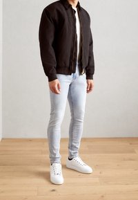 Hombre con chaqueta negra con cremallera, camiseta blanca, vaqueros ajustados de color azul claro y zapatillas blancas de pie sobre un suelo de madera.