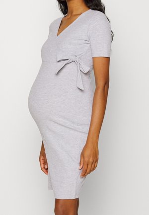 Gravid kvinde iført en tætsiddende, lysegrå, ribstrikket kjole med korte ærmer og et bindebånd i siden ved taljen.