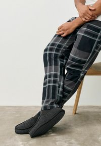 Pantaloni a quadri neri e grigi con una vestibilità ampia, abbinati a pantofole nere con texture, dotate di suola piatta e punta arrotondata.