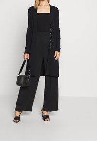Schwarze gerippte Strickjacke mit goldenen Knöpfen, getragen über einem schwarzen Crop-Top und weiter geschnittenen Hosen. Kombiniert mit schwarzen Rutsch-Sandalen und einer kleinen Tasche.