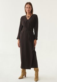 Langes, dunkelbraunes Wickelkleid mit tiefem V-Ausschnitt und langen Ärmeln. Ein passender Gürtel betont die Taille. Kombiniert mit beigen, knielangen Stiefeln.