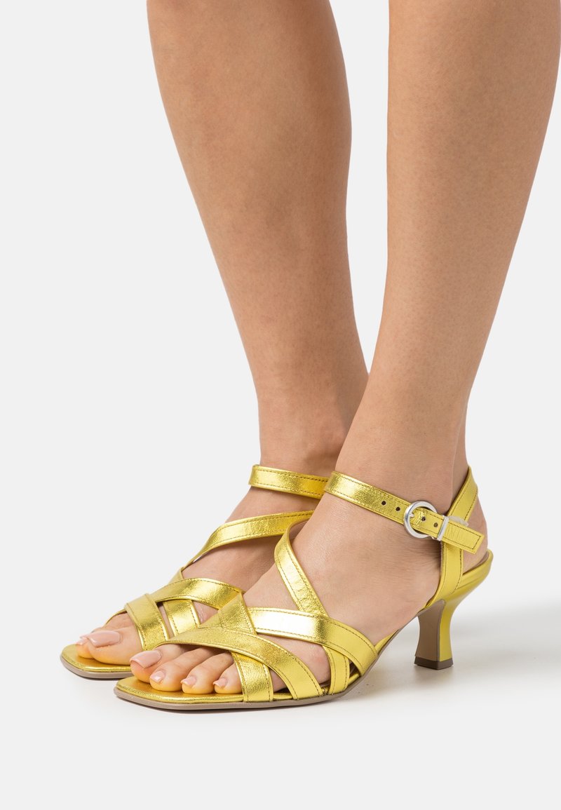 Sandales à talons hauts métalliques jaunes avec des brides croisées et une boucle à la cheville. Conception à bout ouvert avec un petit talon effilé.
