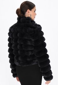 Donna con capelli scuri raccolti a chignon, che indossa grandi orecchini a cerchio e un giubbotto di pelliccia nera, morbido e trapuntato orizzontalmente, di profilo su sfondo bianco.