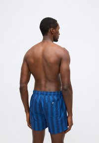 Calções de banho masculinos em azul vibrante com padrão de riscas verticais, cintura elástica e um único bolso traseiro com um pequeno detalhe em forma de logotipo.