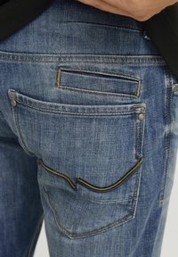Denim traperice u srednje plavoj boji s izblijedjelom teksturom. Imaju dva stražnja džepa, jedan u horizontalnom dizajnu, s kontrastnim šavovima.