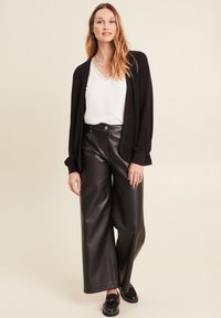 Cardigan long noir, blouse blanche avec dentelle, pantalon large en simili cuir noir et mocassins noirs. Texture lisse et coupe décontractée.