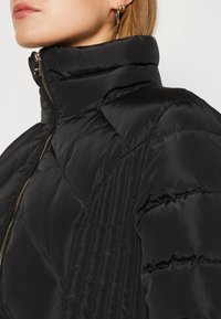 Czarna kurtka puchowa z wysokim kołnierzem, zapięciem na zamek i pikowaną teksturą z poziomymi i pionowymi liniami. Gładki, błyszczący materiał.