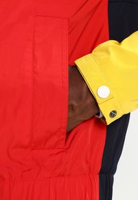 Main à peau foncée insérée dans la poche d'une veste rouge, portant une manche jaune avec un poignet boutonné sur un fond sombre.