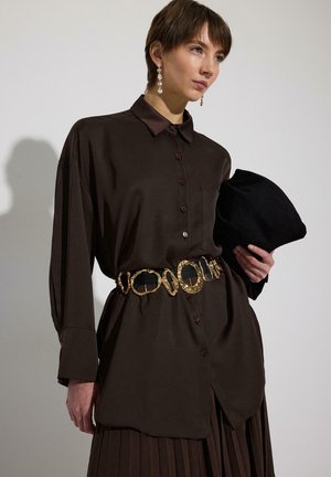 Ženska, oblečena v dolgo rjavo srajco z dolgimi rokavi in zlatim izrazitim pasom, drži črno zloženo torbico na enobarvnem ozadju.