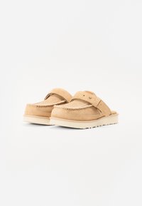 Chaussures à glissière en daim beige avec des détails cousus, une large sangle et une semelle en caoutchouc léger, présentant une finition texturée et un design minimaliste.