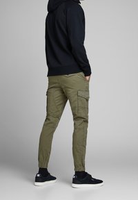 Homme portant un pantalon cargo vert olive, un sweat à capuche noir, des chaussettes noires et des baskets noires, debout les mains dans les poches, de dos.