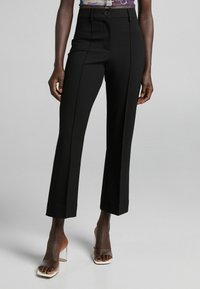 Slim-fit černé kalhoty s předními švy, které nosí stojící osoba v jasných vysokých podpatcích na jednolitém pozadí.