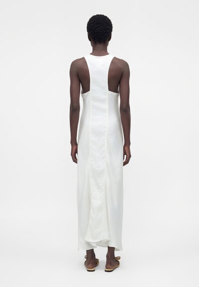 Άτομο με κοντά σγουρά μαλλιά που φορά ένα λευκό αμάνικο μακρύ φόρεμα με εφαρμοστή πλάτη και ανοιχτές πλευρικές επιφάνειες, στέκεται μπροστά σε ένα απλό φόντο.