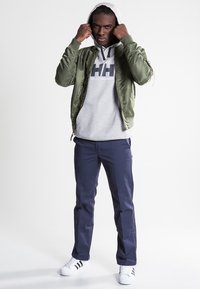Chaqueta bomber de satén verde, sudadera gris con logo, pantalones azul marino y zapatillas blancas. El modelo se encuentra de pie con confianza, con las manos sobre la sudadera.