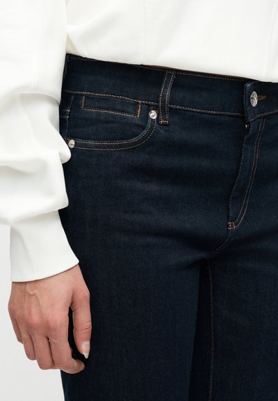 Χέρι ακουμπισμένο στο πλευρό, φοράει σκούρο μπλε τζιν με χάλκινη ραφή και λευκή μπλούζα με μακριά μανίκια και ριμπ μανσέτα.