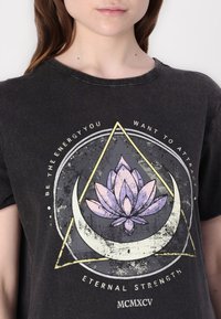 Black cotton T-shirt featuring a lotus flower graphic in purple and white, framed by geometric shapes and text, with a distressed texture.