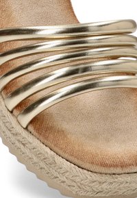 Tirantes metálicos dorados con una suave base de ante beige y una suela de yute trenzada. El diseño presenta cinco bandas elásticas paralelas para mayor comodidad.